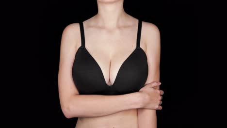 close up of the naked torso of a slim woman with big breasts in a black bra, on a black background in the studio