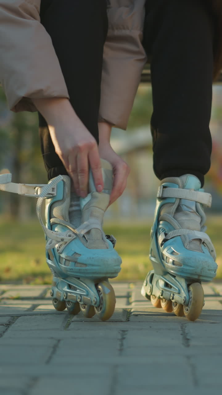 A close-up shot of a person sitting on a park bench, fastening the straps of a rollerblade on one foot while the other foot is already in a rollerblade. The person is not fully visible