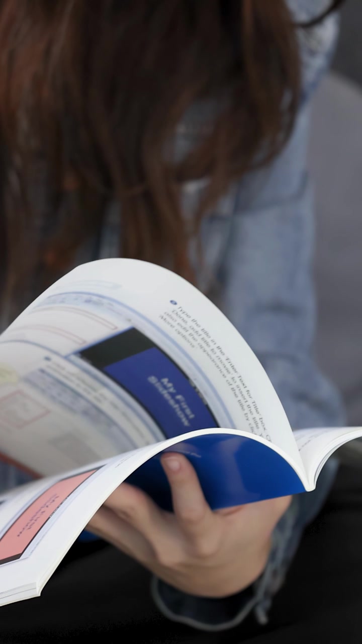 A woman wearing a denim jacket sits indoors while browsing and turning magazine pages slowly