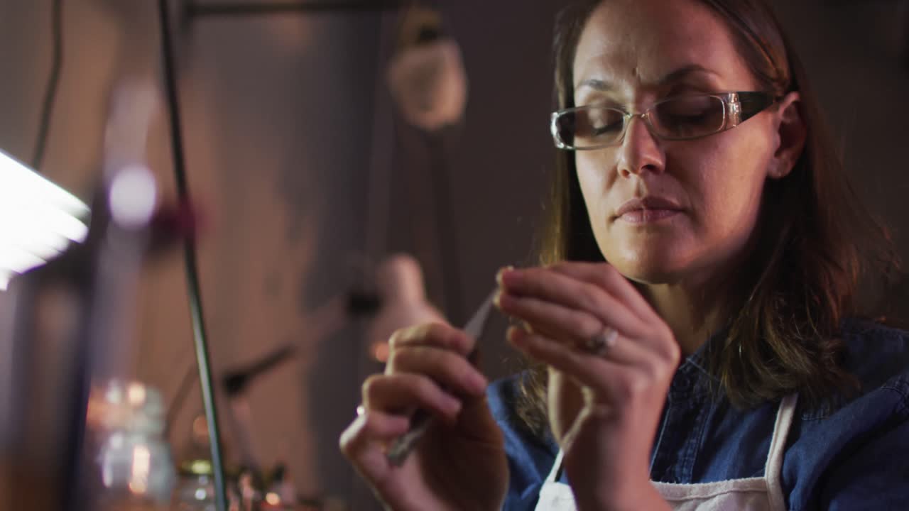 Portrait of caucasian female jeweller wearing glasses, making jewelry and smiling