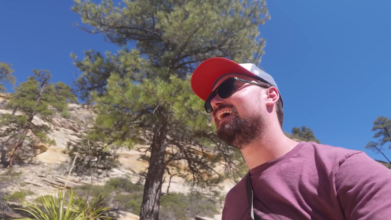A happy man hikes on a mountainous trail with trees, while smiling. He wears a baseball hat and sunglasses on the sunny hike.
