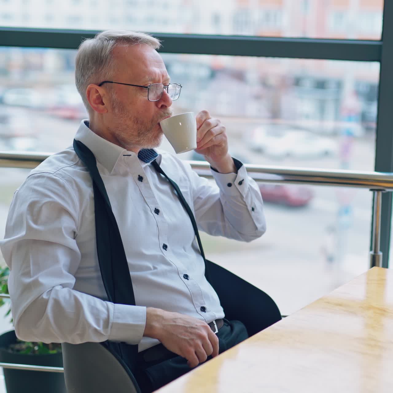 Senior businessman wears white shirt and drinking coffee near big window with city view . Video from the side.