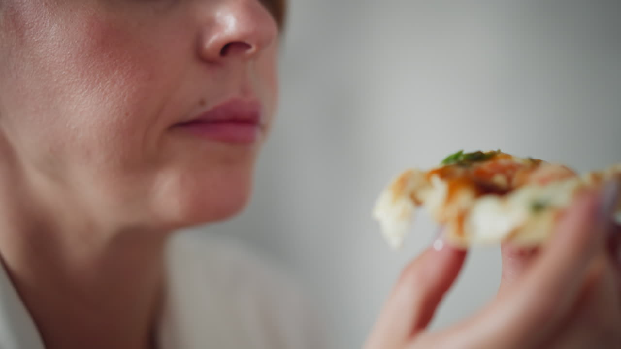 Close up of woman savoring cheesy pizza slice with fresh herbs and sauce, holding delicious food close to mouth, enjoying meal during break, focus on hand and pizza, background softly blurred
