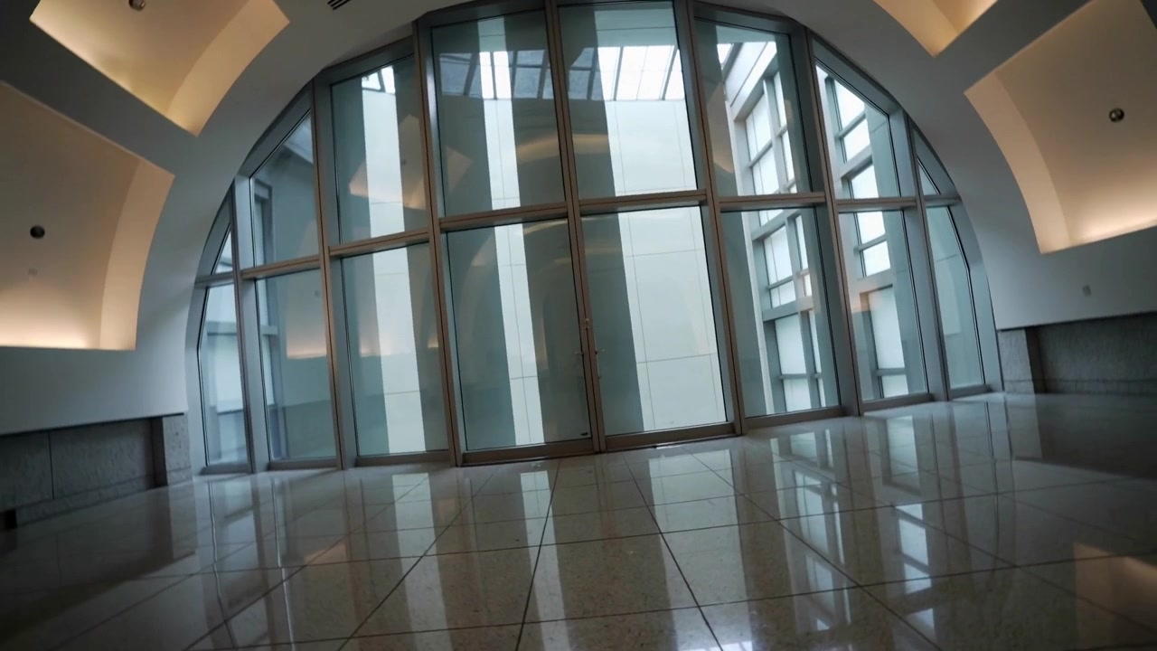 Câmera giratória alinhando parede de vidro arqueada no saguão, revelando portas centrais e reflexo no chão.