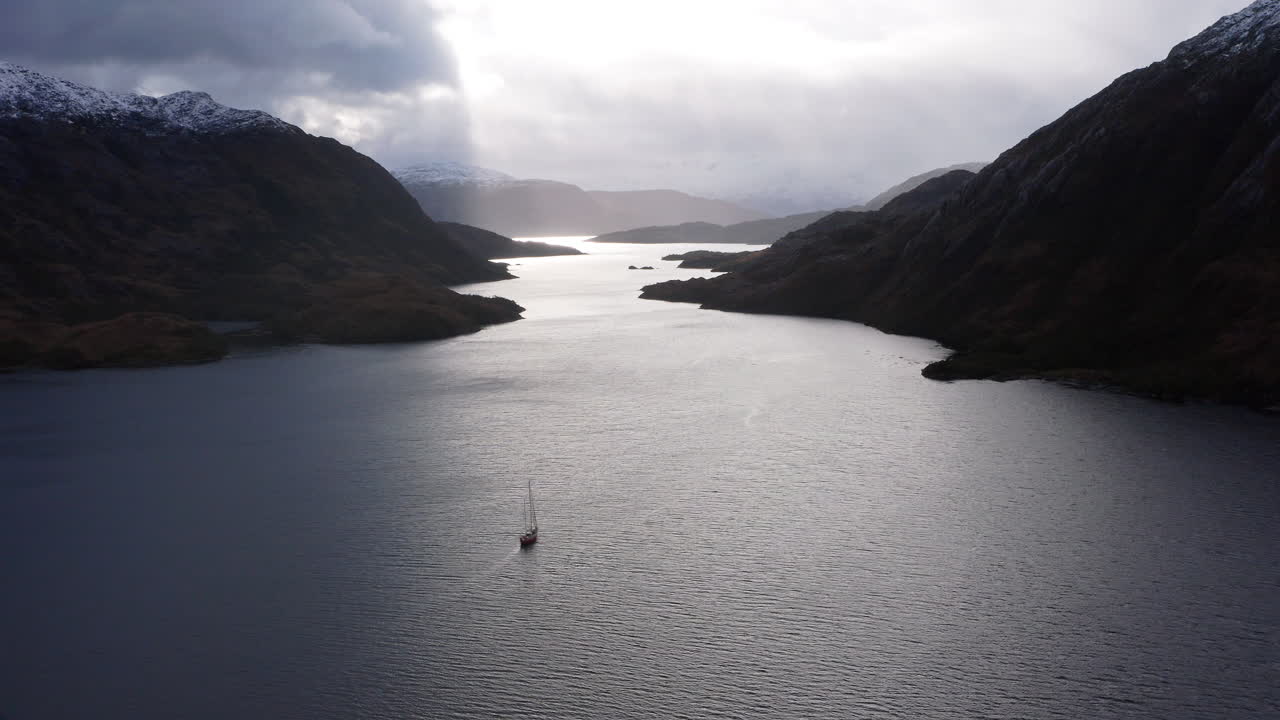 Sailing boat glides down narrow Patagonian fjord under evening shadows and open sky, silhouette, aerial pullback
