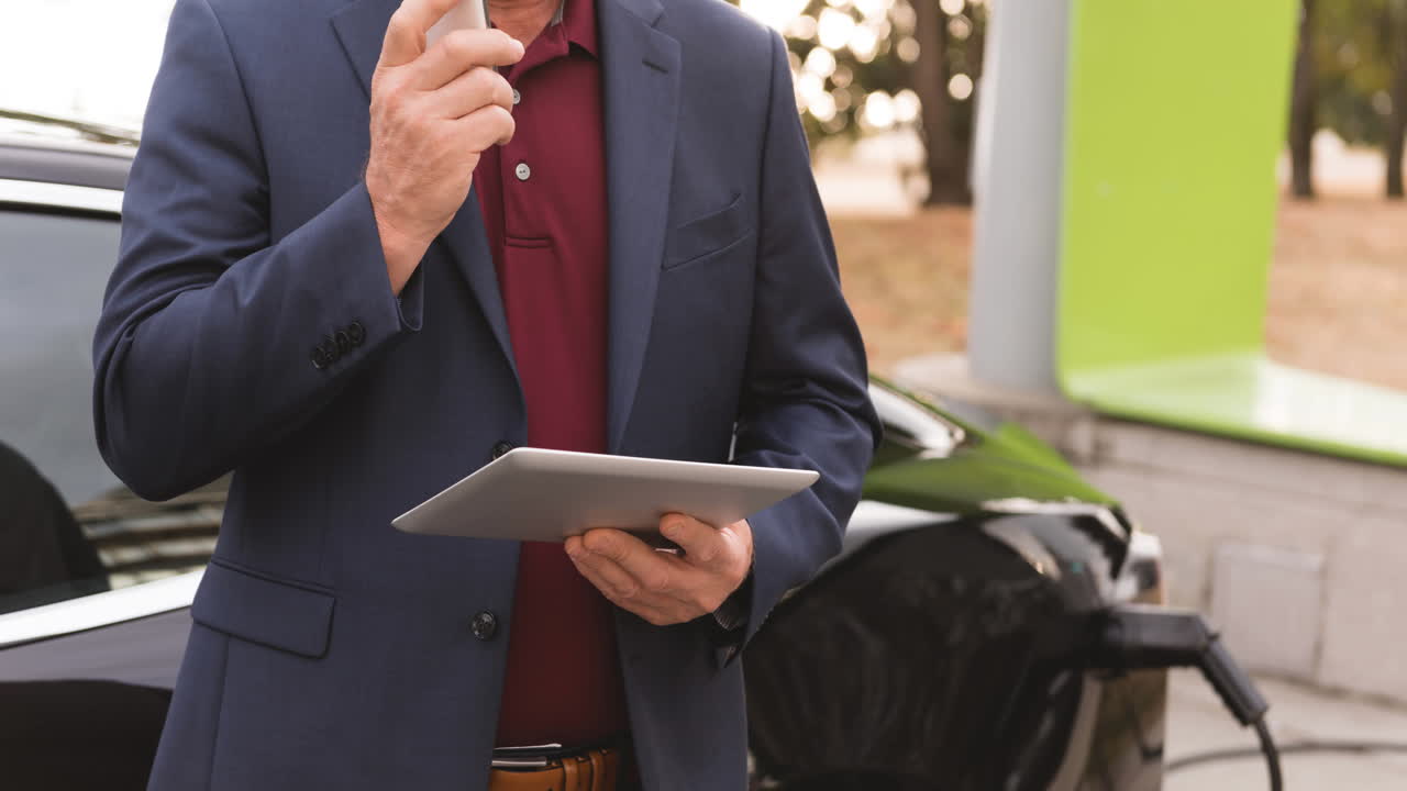sección media de hombre caucásico usando tableta y cargando coche eléctrico