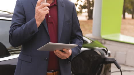 sección media de hombre caucásico usando tableta y cargando coche eléctrico