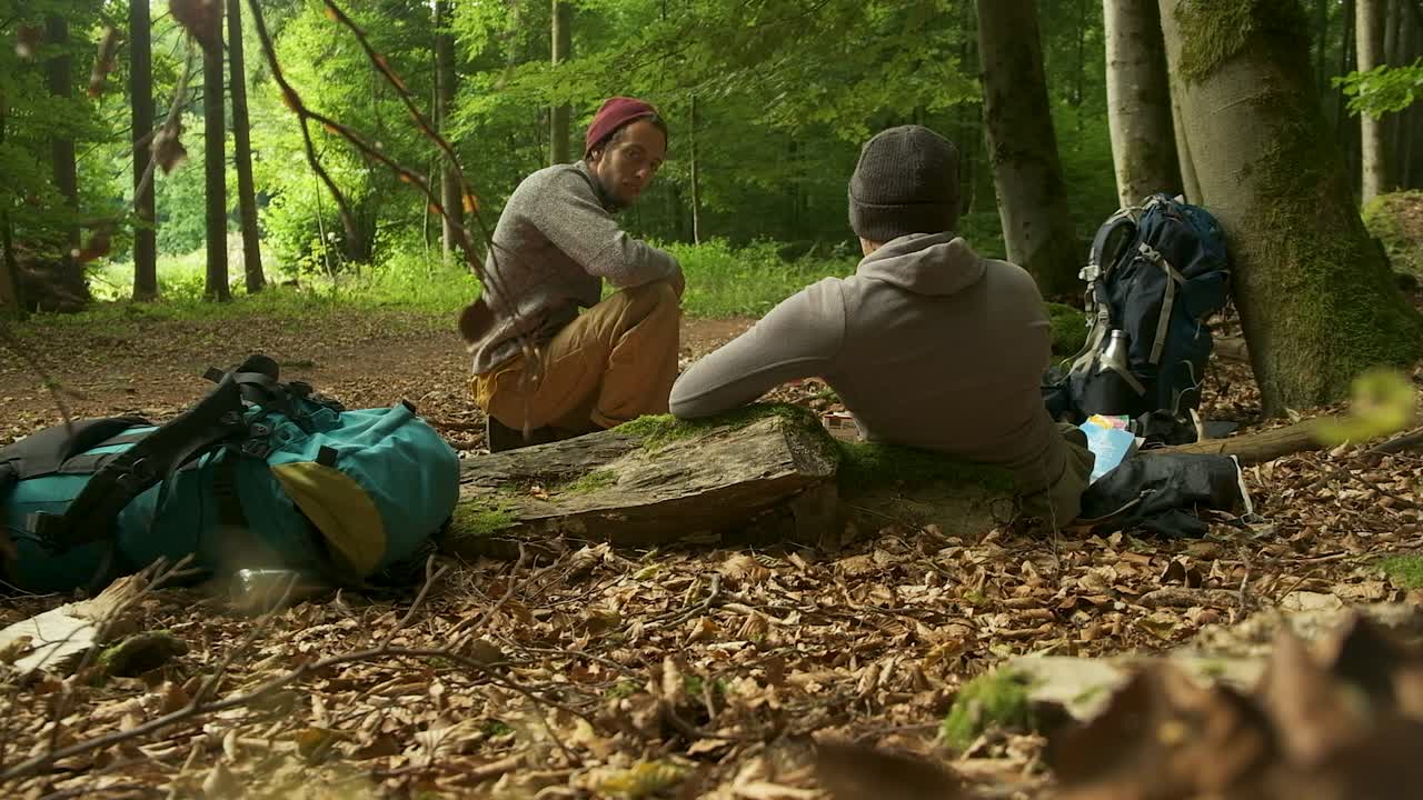 Two friends take a well-deserved rest while hiking in a lush green forest. They sit on the ground and a mossy rock, talking, surrounded by their backpacks