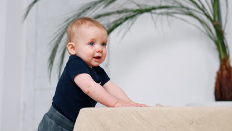 lovely infant boy with red hair stands holding by the sofa. cute little baby portrait.