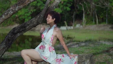 young sexy latina sits on a rock in a long flower dress at the park with a lake in the background on a sunny day
