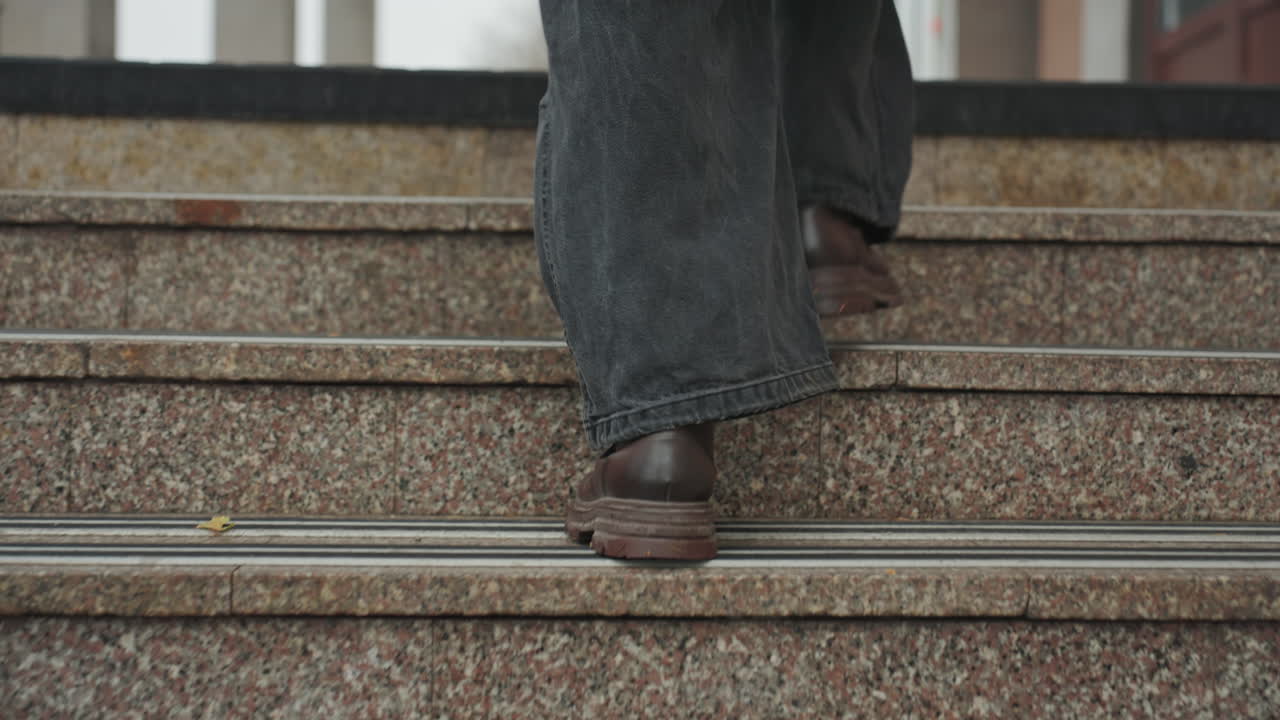 Closeup leather boots ascending stairs, creator climbing municipal steps, damp treads on granite risers, rainy pavement splash, purposeful commute, jeans sway with stride, urban entrance scene