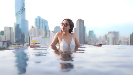 with a background of a modern urban skyline, a pretty young woman leans against the edge of a resort rooftop infinity pool