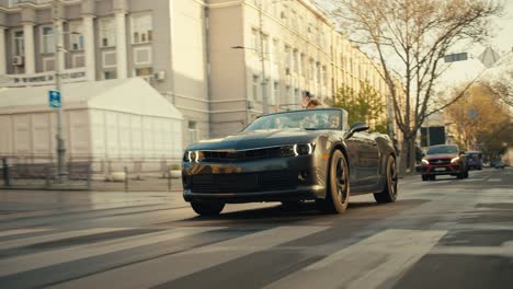 A-dark-gray-Convertible-rides-down-the-street,-a-girl-who-sits-in-the-back-seat-in-a-denim-jacket-waves-her-hands-and-looks-out-of-the-Convertible-in-black-sunglasses