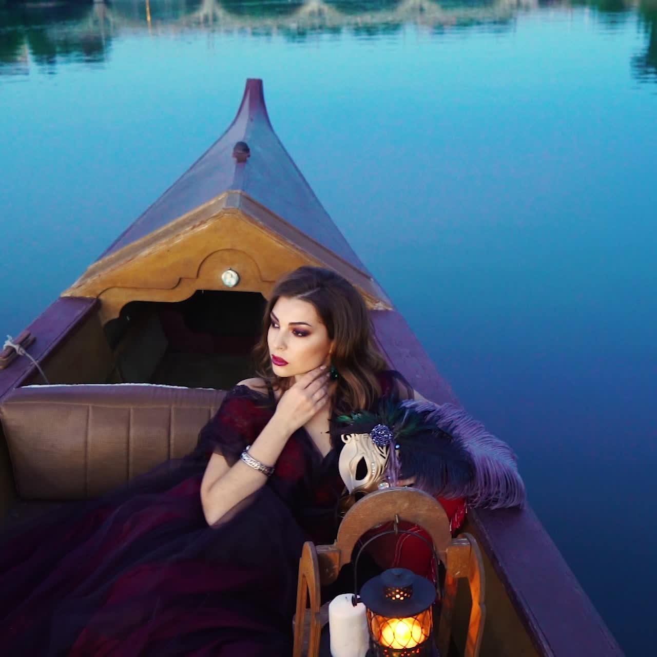 Gorgeous woman in long dark red dress sailing in a boat and touching her earing on blue water background. Beautiful model posing sitting in the gondola in the evening