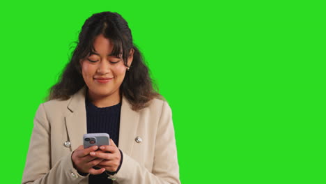 Close-Up-Studio-Shot-Of-Smiling-Businesswoman-Using-Mobile-Phone-To-Message-Or-Browse-Online-Standing-Against-Green-Screen