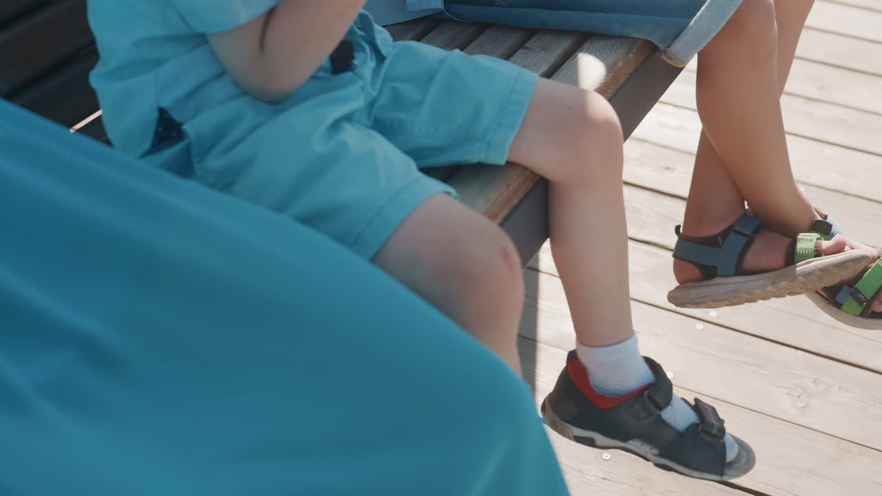 Piedi di bambini e sandali su un ponte illuminato dal sole. Due bambini seduti su una panchina con scarpe da ginnastica e sandali misti, calzini bianchi e movimenti irrequieti giocosi. Ombre calde si estendono sulle assi di legno, in una rilassante gita in famiglia nel fine settimana.