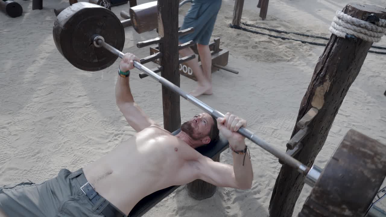 A man working out on a bench press at a beachside fitness area in Koh Phangan, Thailand. The tropical setting combines fitness and beach culture, embodying strength and outdoor exercise.