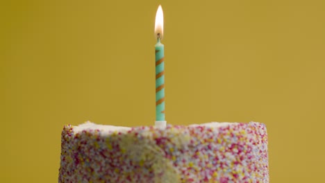 lit single candle on studio shot of revolving birthday cake covered with decorations being blown out