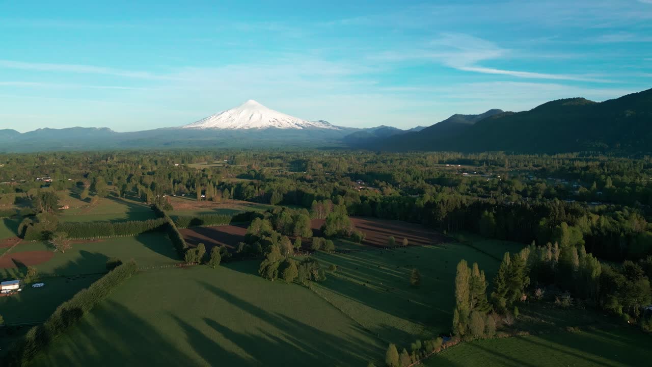 Panoramica aerea del vulcano Villarrica sopra i terreni agricoli e le foreste verdi vicino a Pucón, nel Cile meridionale, con una cima innevata sotto un cielo azzurro.