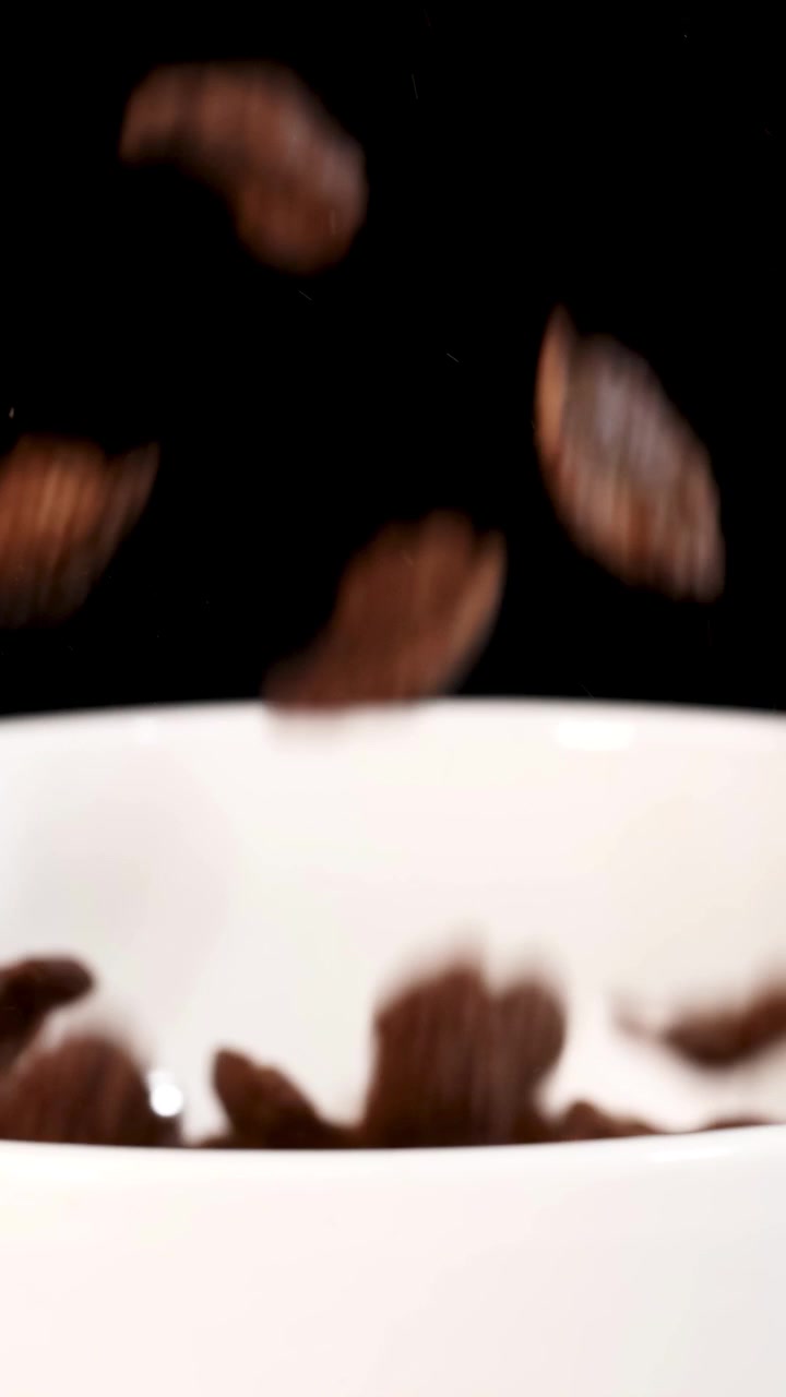 Pezzi di cereali al cioccolato cadono a cascata in una ciotola bianca su uno sfondo nero, creando un'immagine dinamica e appetitosa.