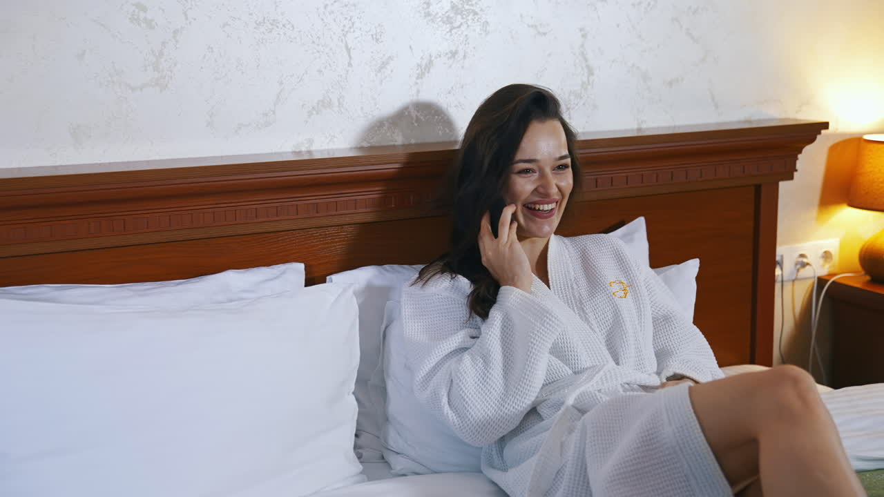 Woman lying in bed at hotel. Happy woman calling on smartphone at hotel room