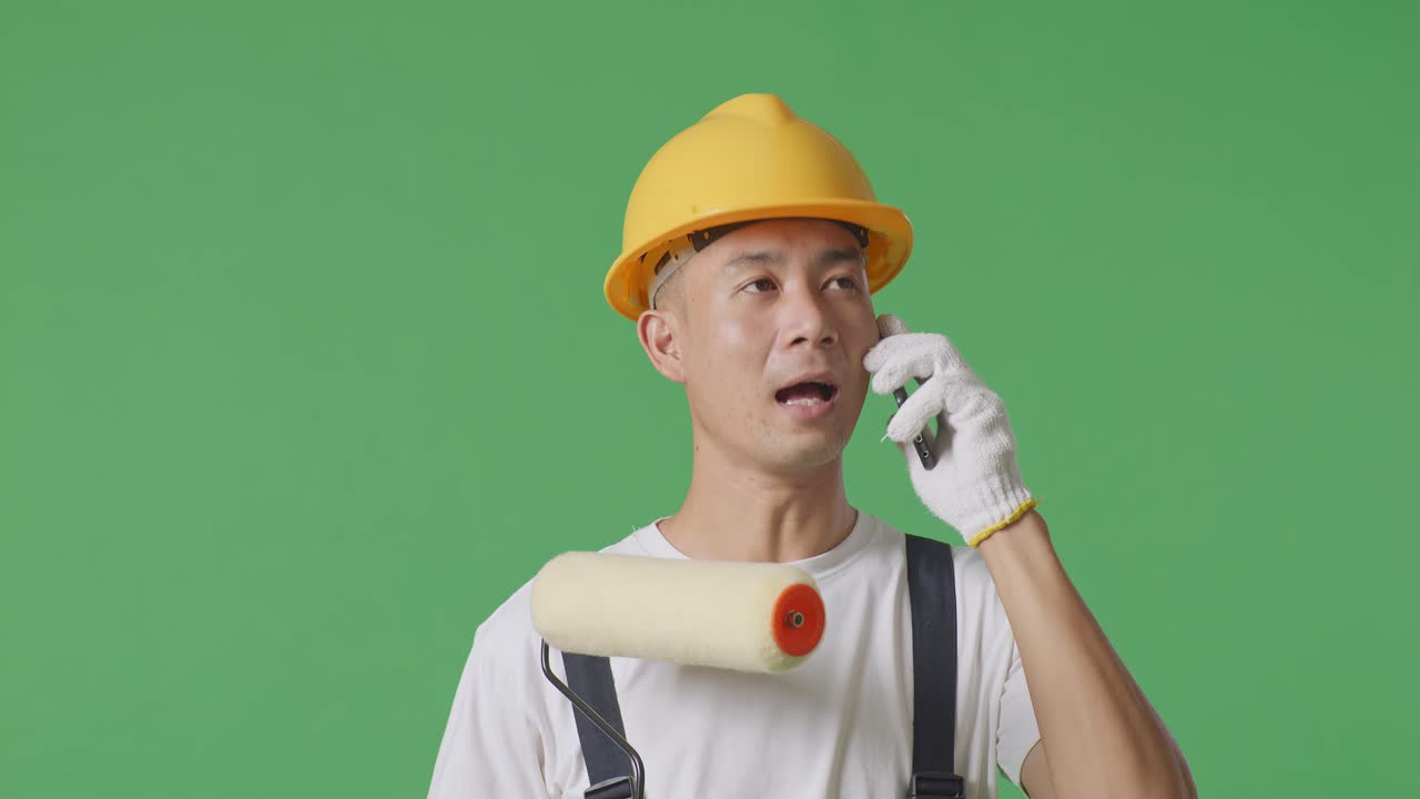 Close Up Of Asian Man Painter Wearing Safety Helmet Talking On Smartphone And Looking Around While Standing In The Green Screen Background Studio