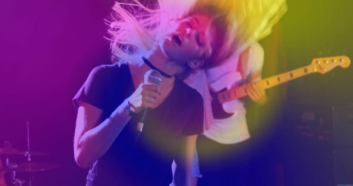 Câmera subindo revelando o vocalista cantando para a multidão, com luzes neon acentuando o microfone, a guitarra e a bateria