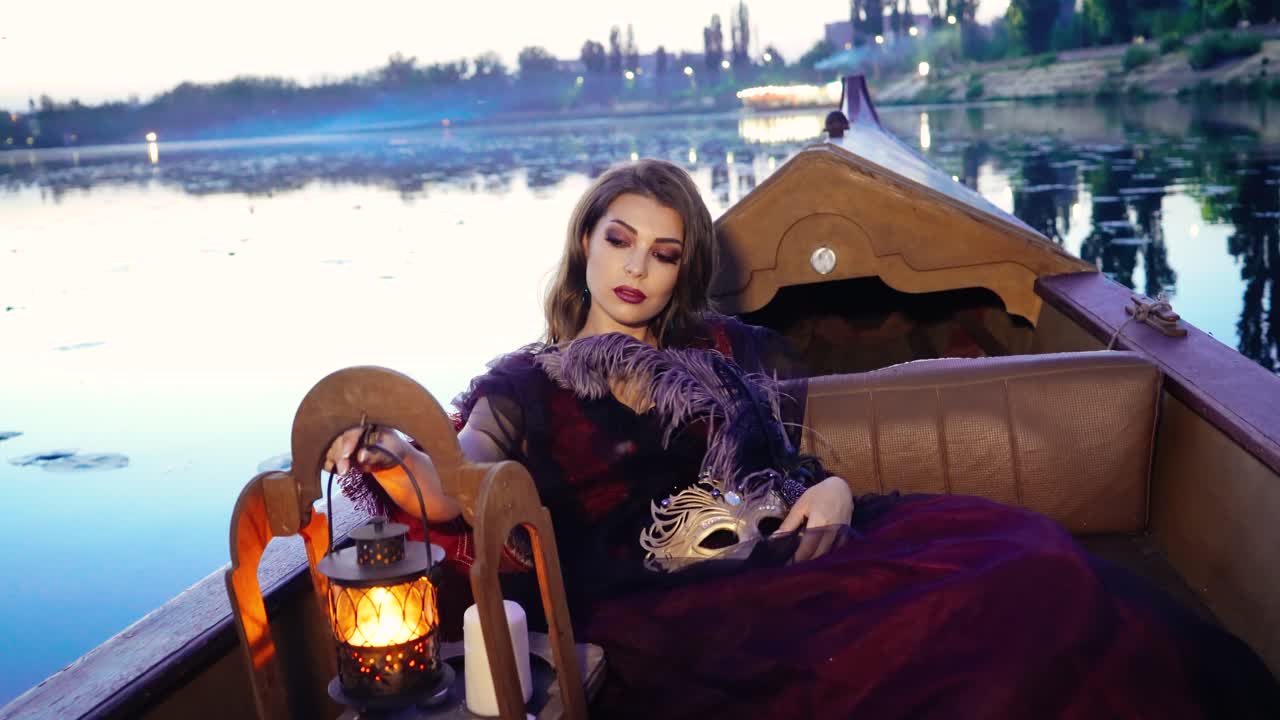 Romantic woman in carnival costume holding feather and mask floating in a boat on the evening river background. Dreamy lady taking a night lamp in her hands at sunset