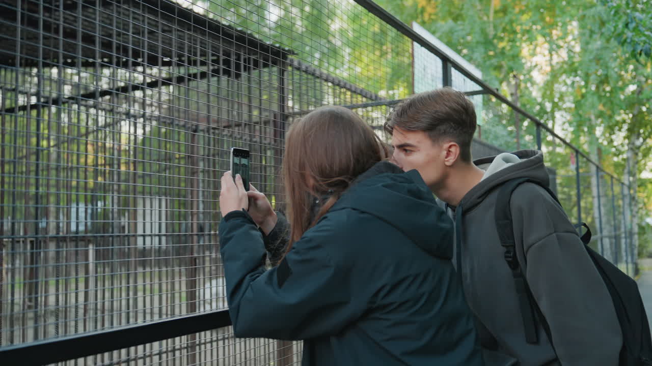 couple photographing through metal enclosure, girl aims smartphone into cage while boy leans over shoulder to peek and whisper, close playful intimacy on park pathway with backpacks and jacketed