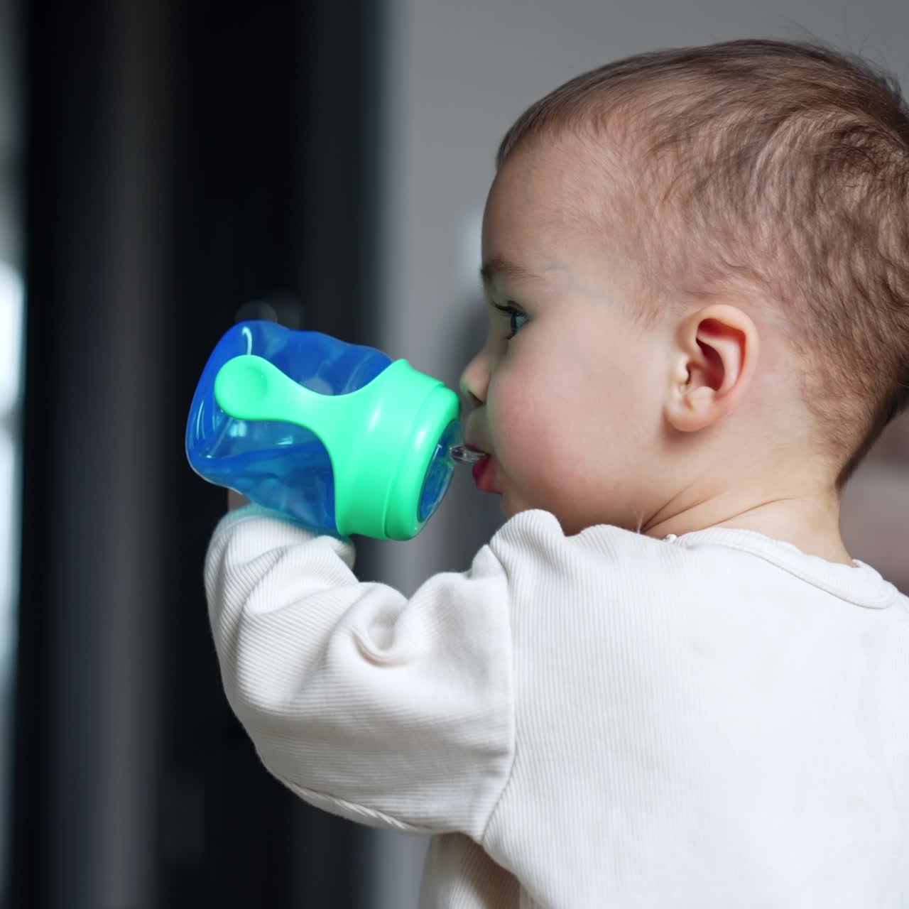 Toddler drinking water from a blue bottle with pacifier. Side view. Baby boy looks up with curiosity. Blurred backdrop