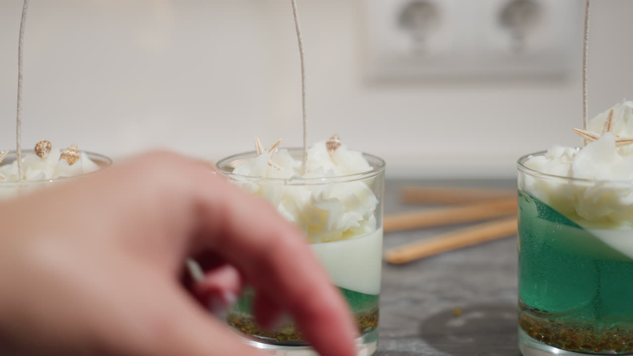 Elegant woman adds brown shell-like decorations to second glass cup filled with whipped white wax topping over layered green and white candle, creating artistic handcrafted centerpiece