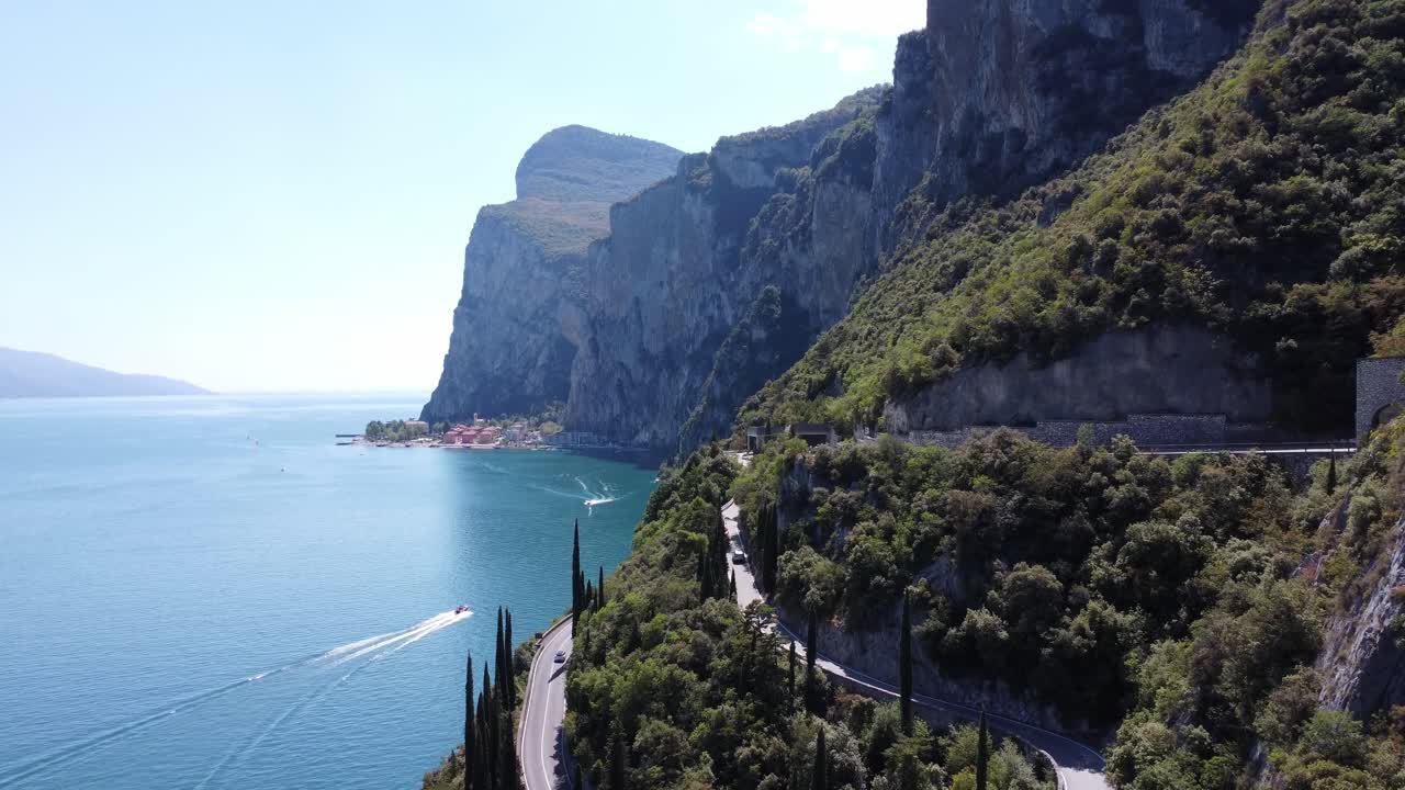 Breathtaking drone shot of Strada della Forra hugging the cliffs above Lake Garda. A speedboat cuts through the blue water while the scenic road winds along the mountain edge.