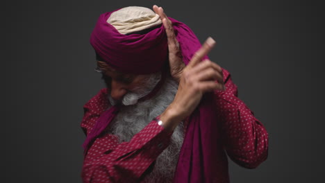 Fotografía-De-Estudio-Con-Iluminación-Tenue-De-Un-Hombre-Sikh-Mayor-Con-Barba-Atando-Una-Tela-Para-Hacer-Un-Turbante-Contra-Un-Fondo-Oscuro,-Filmada-En-Tiempo-Real