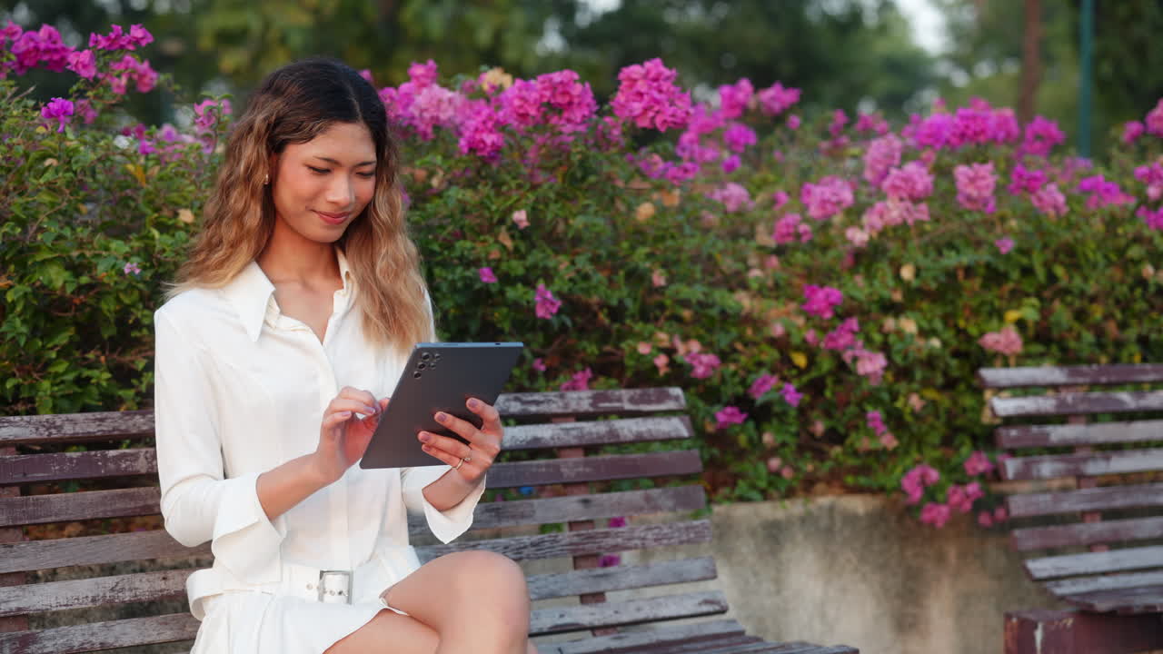 A woman sits on a park bench, using a tablet amidst vibrant pink flowers