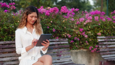a woman sits on a park bench, using a tablet amidst vibrant pink flowers
