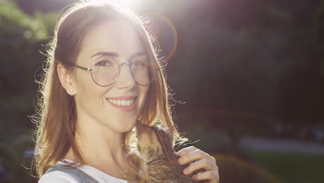 Close-up-view-of-a-caucasian-woman-in-glasses-holding-and-petting-kitty-cat-while-smiling-at-camera-in-the-park-on-a-summer-day