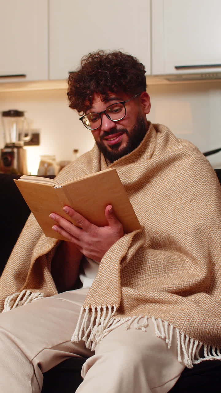 Un homme arabe portant des lunettes, enveloppé dans une couverture, lit un roman lors d’une soirée tranquille, dans une ambiance détendue, concentrée, calme et paisible.