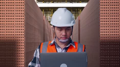 close up of asian male engineer with safety helmet working on a laptop while standing with red brick packed in stacks are stored