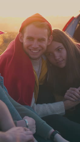 happy young people spend time talking at camp bonfire on meadow in warm autumn evening at sunset close view