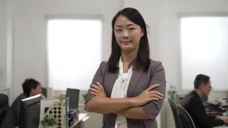 portrait of confident asian businesswoman wearing suit and looking at camera with smiling face and folded arms at bright workplace with coworkers at background