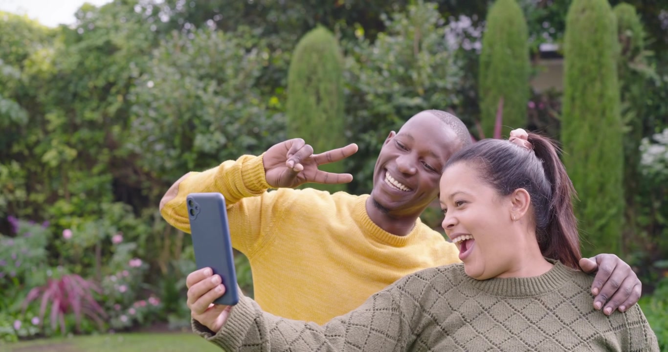 Casal afro-americano no jardim, mulher mexendo no smartphone, homem posando para selfie.