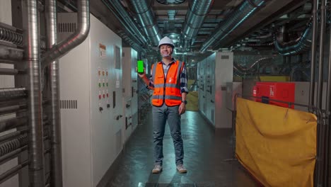 full body of asian male engineer with safety helmet smiling and showing green screen smartphone to the camera while standing in engine control room, work of electrical generators