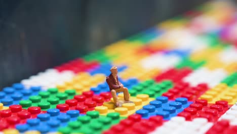 a very small figure depicting a man sitting down is seen placed on top of a colorful lego bricks game