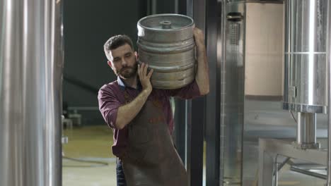 young brewer wearing a leather apron carries a barrel of craft beer at a modern brewery