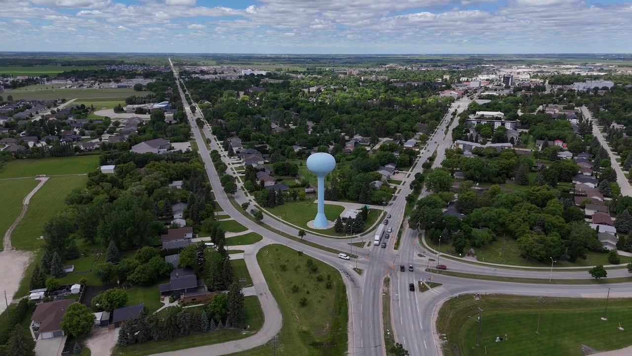 Drone bevæger sig baglæns væk fra et lyseblåt vandtårn i Steinbach, Manitoba, og afslører den hævede tank ved siden af veje, biler, huse, træer, forstæder, træalléer og grønt græs om sommeren.
