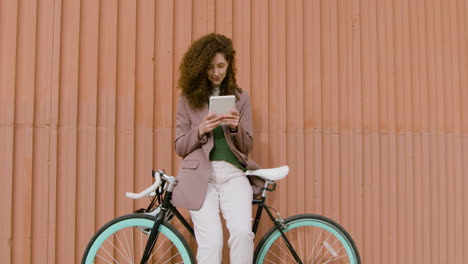 mujer rizada con ropa formal usando una tableta mientras se apoya en bicicleta frente a un edificio de metal prefabricado