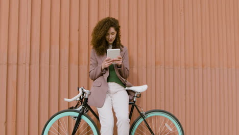 curly woman in formal clothes using a tablet while leaning on bike in front of a prefab metal building