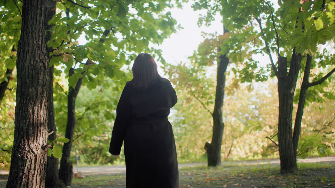 Upward view of cozy young lady strolling through autumn forest, hand relaxed by side, sipping warm drink, long coat and boots moving along leafy path, soft sunlight filtering through canopy
