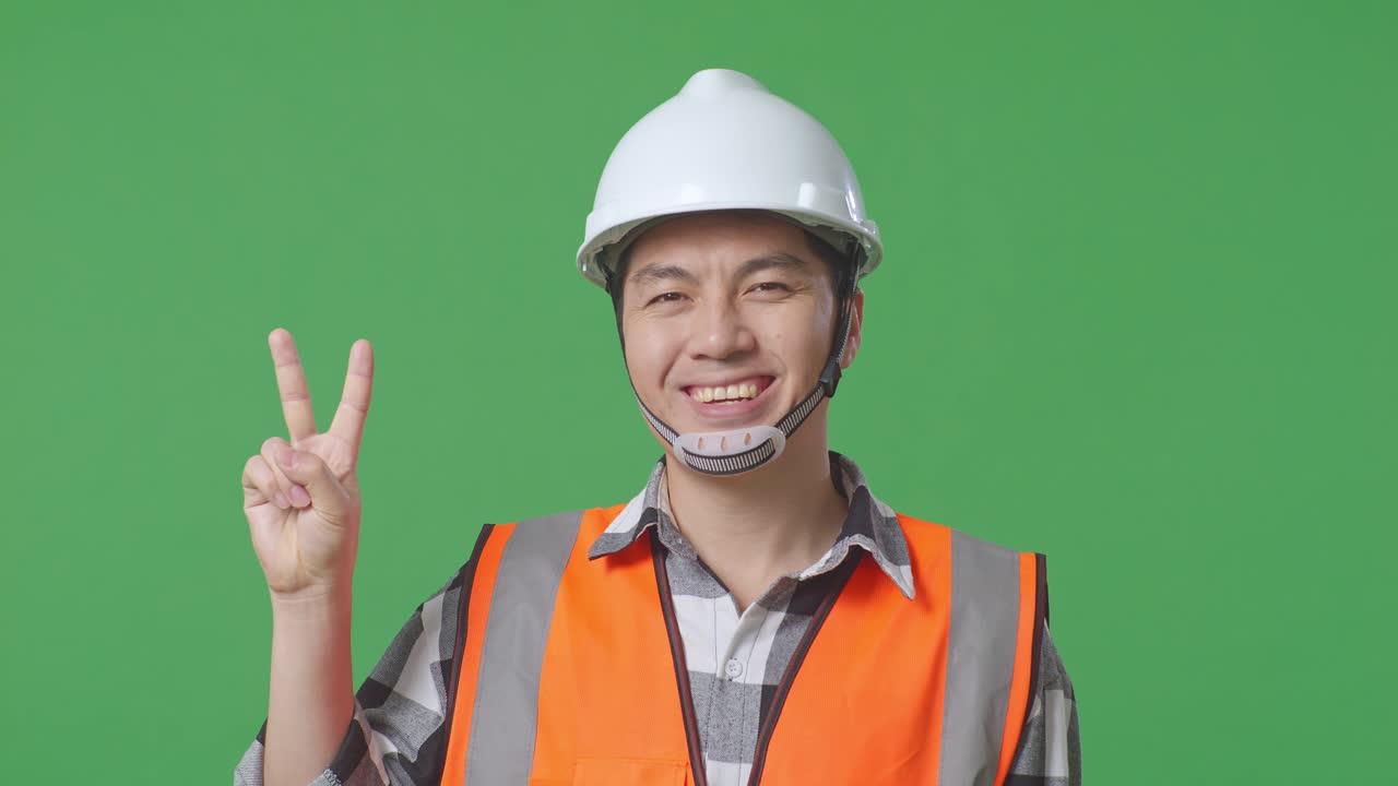 Close Up Of Asian Male Engineer With Safety Helmet Smiling And Showing Peace Gesture While Standing In The Green Screen Background Studio