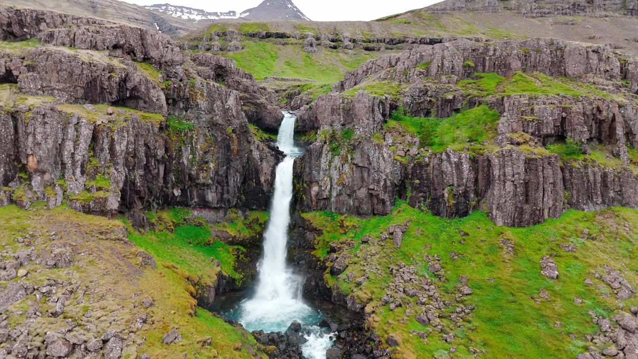 Et fantastisk luftfoto af Snædalsfoss vandfaldet på Island, der viser hvidt vand, der fosser ned over mørke basaltklipper, omgivet af grønne mosklædte bakker. Ideel til rejse- og naturdokumentarer.